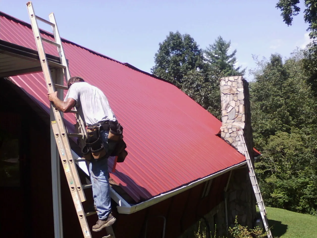 Roofer on a ladder beside a red metal roof and stone chimney — Soffit & Fascia Repair by Roofing Brothers in Jacksboro